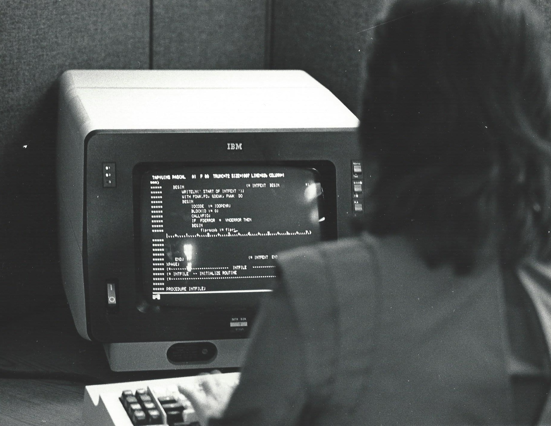 An Informatics General computer programmer working in a cubicle at the company's New York City office. The programmer is looking at Pascal language code for the TAPS product on an IBM 3270 terminal. Date: October 1, 1983
